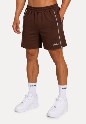 Persona muscolosa che indossa pantaloncini marroni con elastico in vita e piping bianco, calzini bianchi con scritte e scarpe da ginnastica bianche con lacci.