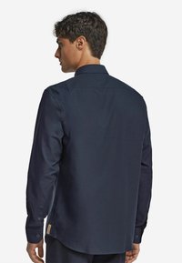 Marinblus med lång ärm som har en klassisk krage, knappmanchetter och rak nederkant. Tillverkad av glatt, texturerat tyg med minimal detaljer.