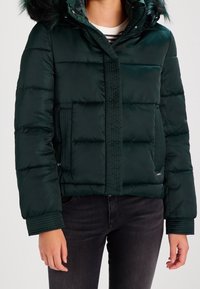 Giacca a vento verde con cappuccio foderato in pelliccia, chiusura con zip e polsini a coste. Presenta cuciture orizzontali e tasche laterali.