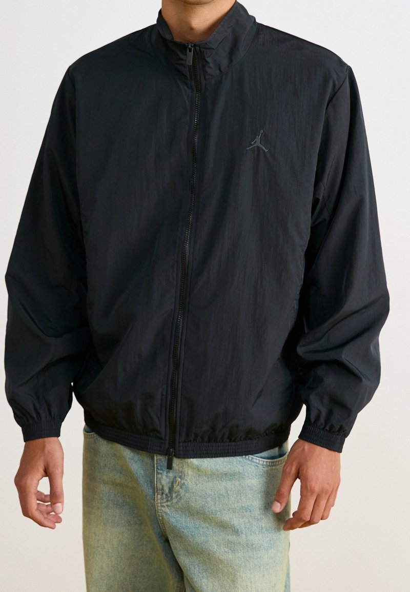 Homme portant une veste noire zippée avec un petit logo sur la poitrine et un jean en denim bleu clair, debout devant un fond uni.