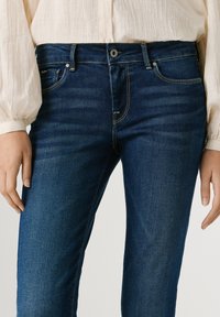 Άτομο που φοράει σκούρο μπλε τζιν παντελόνι και λευκό μπλουζάκι με μακριά μανίκια, όρθιο με τα χέρια χαλαρά δίπλα στο σώμα.