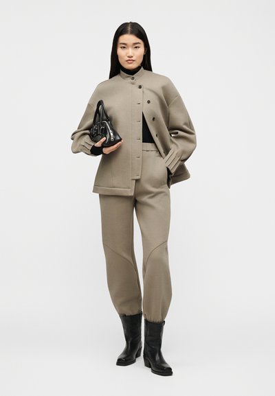 Γυναίκα που στέκεται φορώντας ταμπά μπουφάν με κουμπιά και αντίστοιχα παντελόνια φόρμας, μαύρο ζιβάγκο, μαύρες μπότες, κρατώντας μικρή μαύρη τσάντα.