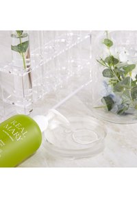 Bottiglia verde con etichetta "REAL MARY", piatto di vetro chiaro, portatubi trasparente e contenitore di vetro con fogliame finto su una superficie chiara.