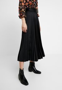 Svart plisserad midikjol med en slät textur, figursydd midja, i kombination med blanka svarta ankelstövlar med spetsig tå och klack i blockformat.