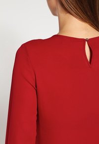 Vêtement rouge avec un détail dos en forme de trou de serrure et des manches longues. Texture lisse et coupe ajustée pour un look sur mesure.
