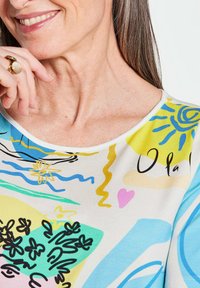 Multicolor kortemouwen top met abstracte vormen, voornamelijk geel, blauw en groen, met een ronde halslijn en een zachte textuur.