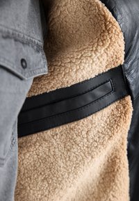 Veste polaire sherpa beige avec accents en cuir noir et partie d'une manche en tissu gris avec un bouton-pression noir visible.