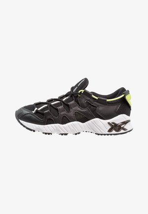 Zapatilla deportiva en blanco y negro con parte superior de malla y cuero, suela blanca gruesa y texturizada, detalles amarillos y cordones negros.