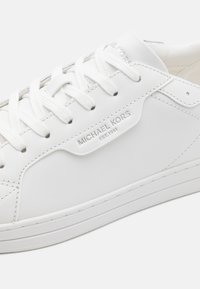 Hvite skinnsko med flate hvite snørebånd, med en sydd design og preget "MICHAEL KORS"-logo på siden. Gummisåle.