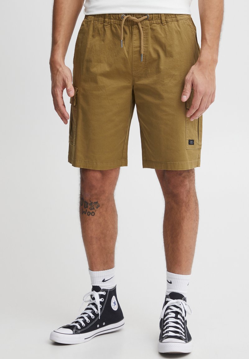 Khaki cargo shorts with an elastic waistband, drawstring closure, side pockets, and a subtle logo patch. Worn with black high-top sneakers.