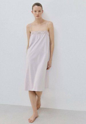 Frau mit nackten Füßen in einem blasslila ärmellosen Midikleid mit Spitzensaum, die vor einer schlichten weißen Wand steht.