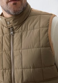 Primo piano di un uomo che indossa un gilet smanicato trapuntato di colore marrone chiaro su una camicia a maniche lunghe beige con texture.