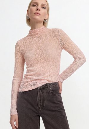 Donna che indossa un top a maniche lunghe texturizzato rosa pallido e jeans scuri a vita alta, in piedi con una mano sui fianchi su sfondo bianco.
