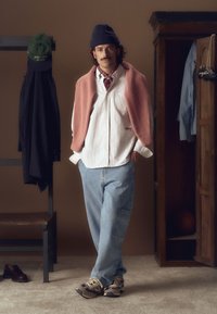 Denim blaue Hose, weiß gestreifte Hemd mit Knöpfen, rosa gestrickter Pullover über die Schultern gelegt, braune und beige Sneakers, trägt eine dunkle Mütze.