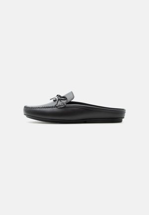 Μαύρο δερμάτινο μοκασίνι χωρίς πλάτη με λεπτομέρεια φιόγκου στην κορυφή, ραμμένα άκρα και επίπεδη σόλα σε μονόχρωμο φόντο.