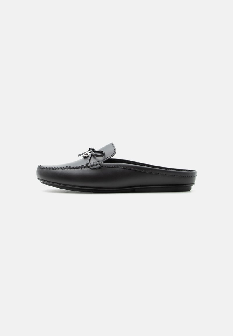 Μαύρο δερμάτινο μοκασίνι χωρίς πλάτη με λεπτομέρεια φιόγκου στην κορυφή, ραμμένα άκρα και επίπεδη σόλα σε μονόχρωμο φόντο.