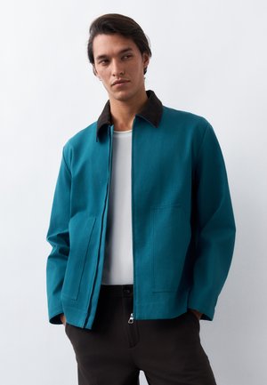 Νέος άνδρας που φοράει πετρόλ ζακέτα με φερμουάρ και μαύρο γιακά πάνω από λευκό πουκάμισο, στέκεται με τα χέρια στις τσέπες σε λευκό φόντο.