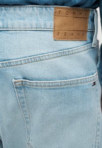 Κοντινό πλάνο πίσω τσέπης από τζιν ανοιχτού μπλε χρώματος με καφέ δερμάτινη ετικέτα που γράφει "Tommy Jeans" και μικρή σημαία της μάρκας στη γωνία της τσέπης.