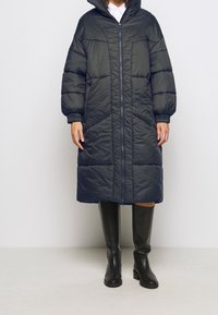 Piumino blu navale con cappuccio, zip frontale a lunghezza completa e polsini elastici. Il design presenta cuciture diagonali e tasche laterali.