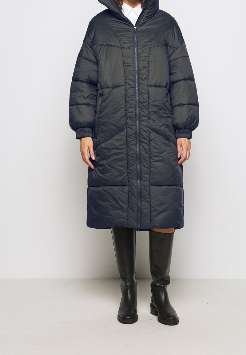 Piumino blu navale con cappuccio, zip frontale a lunghezza completa e polsini elastici. Il design presenta cuciture diagonali e tasche laterali.