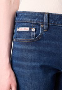 Fatos de ganga azul escuro com costura laranja, apresentando um bolso frontal com um rótulo visível da Calvin Klein e fecho de pressão em metal.