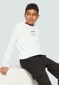 Jongen met krullend haar, gekleed in een witte lange mouwen shirt met tekst en zwarte cargobroek, zittend op een witte blok, kijkend opzij.
