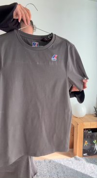 Personne tenant un t-shirt K-Way gris foncé sur un cintre à l’intérieur, près d’une table en bois avec une lampe ronde blanche et une boîte décorative.