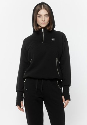 Kobieta ubrana w czarną bluzę z kapturem i dopasowane czarne spodnie dresowe z srebrnymi suwakami, stojąca na jednolitym białym tle.