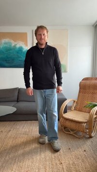 Maglione nero con zip, jeans dritti azzurri, scarpe grigie. In piedi in una stanza con un divano grigio e una sedia in rattan.