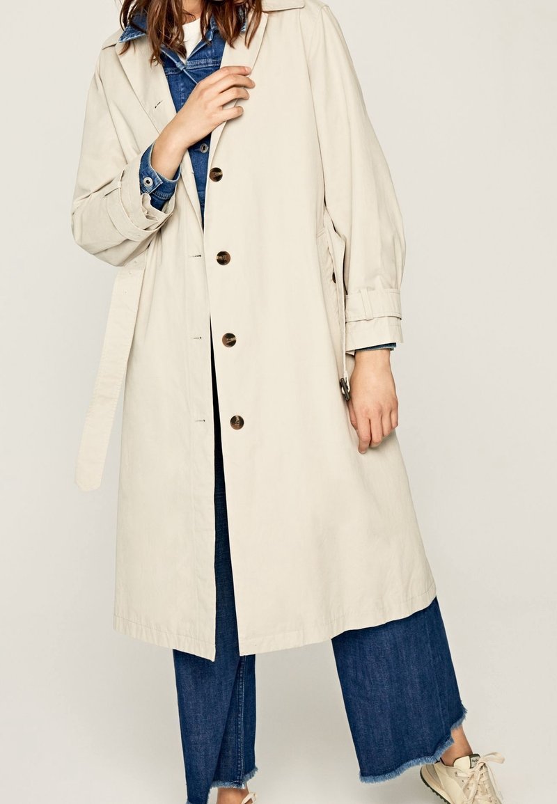 Trench beige avec une fermeture à boutons, des poches latérales et une ceinture. Porté sur une chemise en jean et un jean à jambes larges. Tissu doux et coupe décontractée.