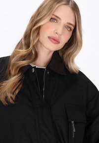 Giacca utilitaria nera con colletto in corduroy marrone, chiusura con zip e dettagli sulle tasche, che mostra una texture liscia e una vestibilità strutturata.
