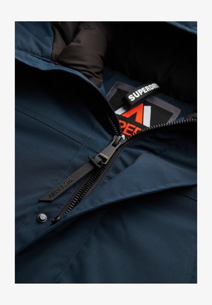 Marineblauwe geïsoleerde jas met een interieur dat een zwarte rits, zwarte voering en een zichtbare Superdry logo-tag bevat. Zachte textuur en stevige uitstraling.