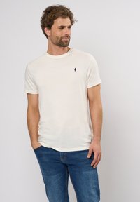 Beige bomull t-skjorte med rund hals og korte ermer. Har en liten marineblå logo på venstre bryst. Stylert med blå jeans.