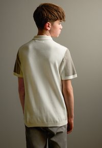 Krēmkrāsas polo krekls ar teksturētām īsām piedurknēm; gluda ķermeņa auduma; ribēti akcenti apkaklē un piedurknēs; pieguļošs dizains redzams no aizmugures.