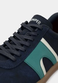 Laivastonsininen tennari mustilla nauhoilla, vihreillä ja valkoisilla nahkaisilla sivuraitojen yksityiskohdilla sekä ruskealla pohjalla, jossa näkyy brändin nimi kielen kohdalla.