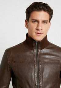 pruun nahktagi ribadega kaelusega, esimese lukuga ja detailse õmblusega. Sile tekstuur, sobitatud disain ja õlavarre aktsendid.