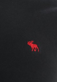 Svart T-shirt med en mjuk textur som har en liten röd broderad älg-logotyp på vänster sida. Enkelt design utan ytterligare mönster.