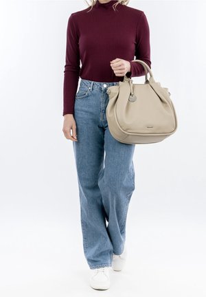 Hellbeige Leder-Handtasche mit runder Form und Griffen oben, gehalten von einer Person, die einen burgunderfarbenen gerippten Rollkragenpullover und blaue Jeans trägt.