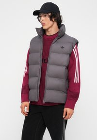 Jovem vestido com um boné preto, colete cinza acolchoado, suéter maroon da Adidas com riscas brancas e calças pretas, contra um fundo branco liso.