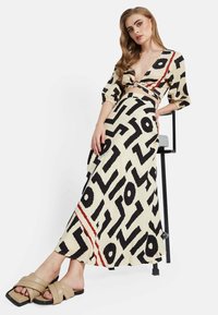 Vestido maxi en crema con patrones abstractos en negro y acentos rojos. Escote en V, mangas abullonadas y detalles cut-out en la cintura. Combinado con sandalias beige.