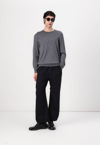 Pull gris à manches longues associé à un pantalon noir à jambes larges. Le mannequin porte des lunettes de soleil noires et des chaussures noires. Design épuré et minimaliste.