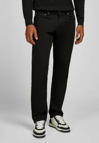 Homme portant un jean noir, un haut à manches longues noir et des baskets noir et blanc, se tenant devant un fond clair.