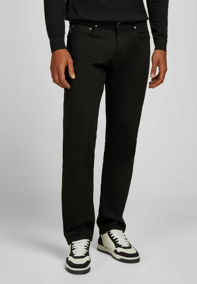 Homme portant un jean noir, un haut à manches longues noir et des baskets noir et blanc, se tenant devant un fond clair.