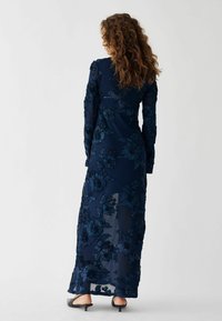 Granatowa sukienka maxi z haftem w kwiaty, z długimi rękawami, przezroczystego materiału i teksturowanymi detalami. Slim fit z delikatnym trenem na dole.
