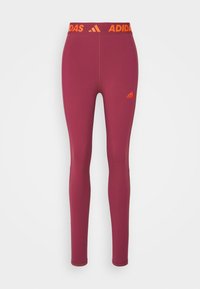 Bordeaux leggings met een oranje elastische tailleband met "ADIDAS" branding. Glad materiaal, strakke pasvorm en taps toelopende benen.