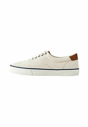 Beige lage sneaker van canvas met een witte rubberen zool, witte veters, marineblauwe streepdetails en een bruine suède hielstuk, zijaanzicht op een witte achtergrond.