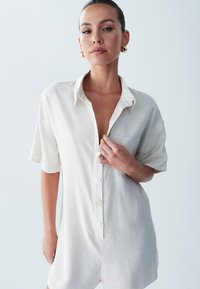 Kratke rokave, svetlo bež kombinezon z gumbom na sprednji strani, ovratnikom in žepom na levi strani. Mehka, teksturirana tkanina.