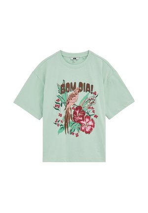 T-shirt vert clair avec motif floral tropical, un perroquet perché et le texte « BOM DIA ! » et « TROPIC CANA » en marron et rouge.