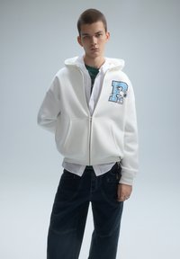 Witte hoodie met ritssluiting, een voorzak en een grafisch ontwerp. Draagt over een groen gestreept shirt, gecombineerd met donker wijde broekspijpen.