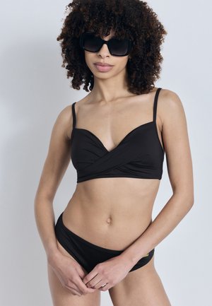 Kvinne med krøllete hår som bærer store svarte solbriller og en svart to-delt badedrakt, poserer foran en ensfarget lys grå bakgrunn.